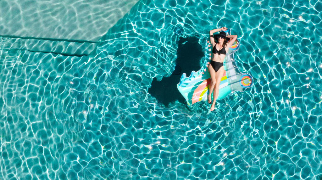 Model Stephanie looks picturesque, smiling softly in a sunhat and black bikini during an aerial pool portrait shoot