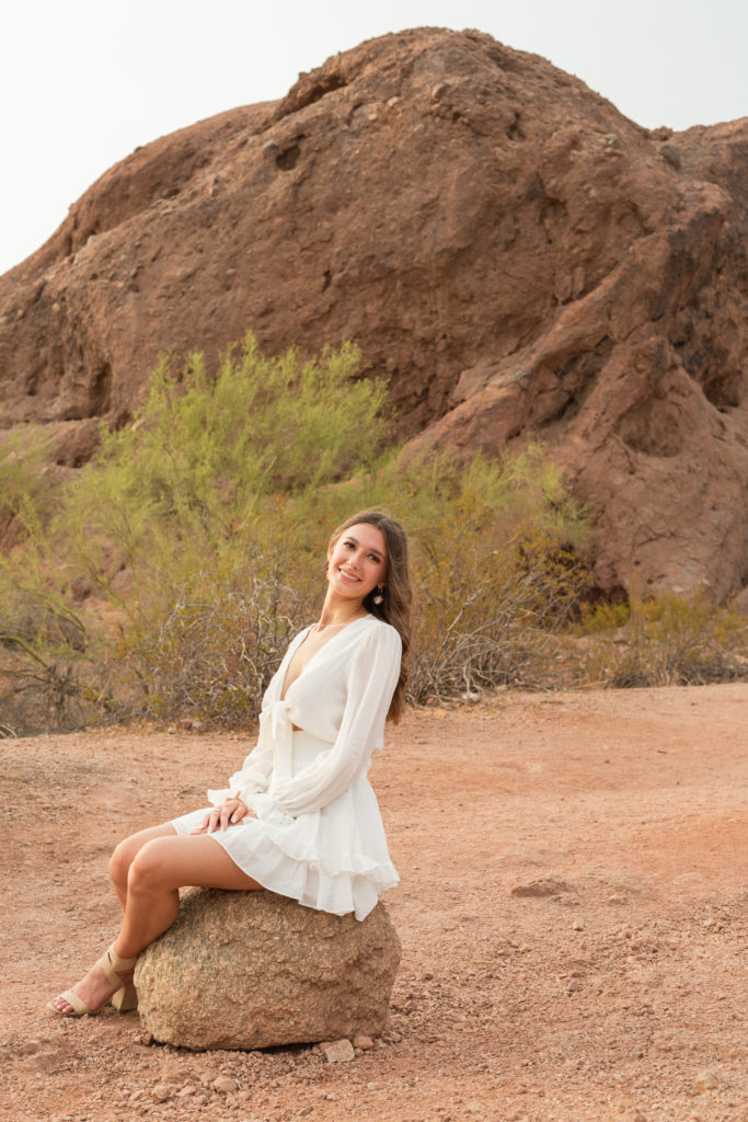 Seated on a round, red rock in Papago Park, 2022 senior grad Kylie leans back smiling during her portrait session