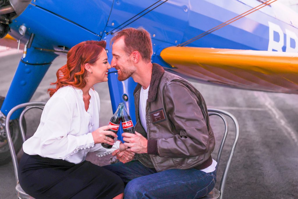 Clients Jennifer and Ben share smiles and a Coca-Cola during an engagement portrait session at an airport in Mesa, Arizona with Bayley Jordan. 