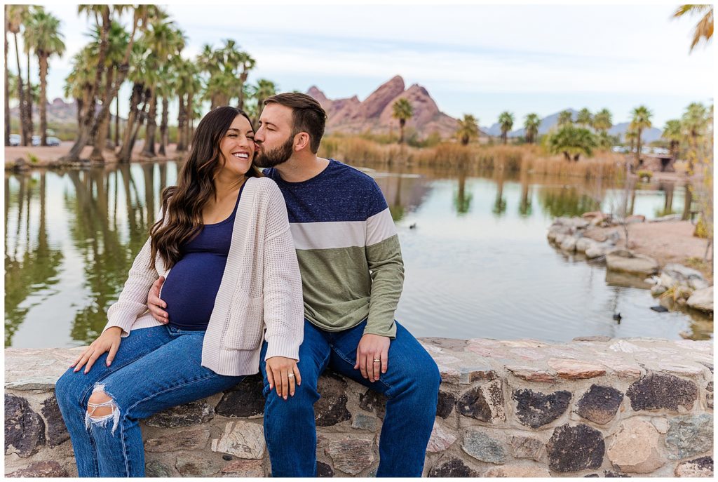 Husband kisses wife on cheek, making her giggle, during romantic maternity portrait session in Tempe, Arizona with Bayley Jordan Photography