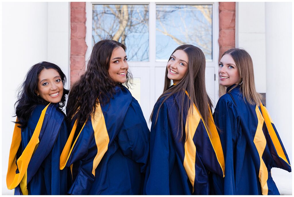 A group of four friends stand in front of a historic building at NAU wearing caps and gowns to celebrate their upcoming graduation
