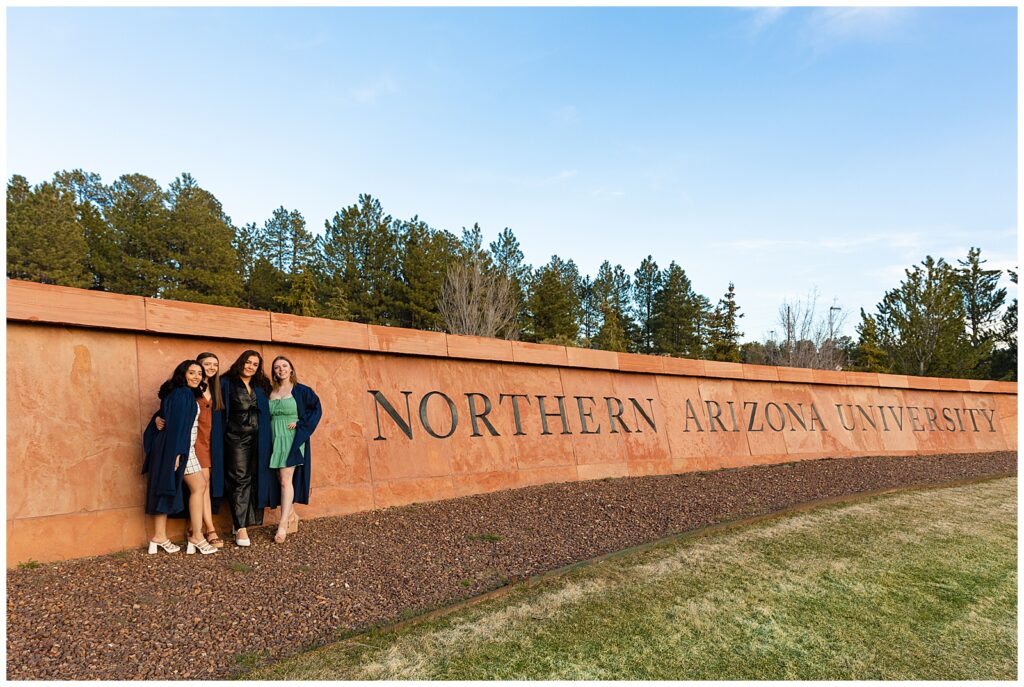 Four Master's graduates pose for portraits together in front of large Northern Arizona University sign at Flagstaff campus