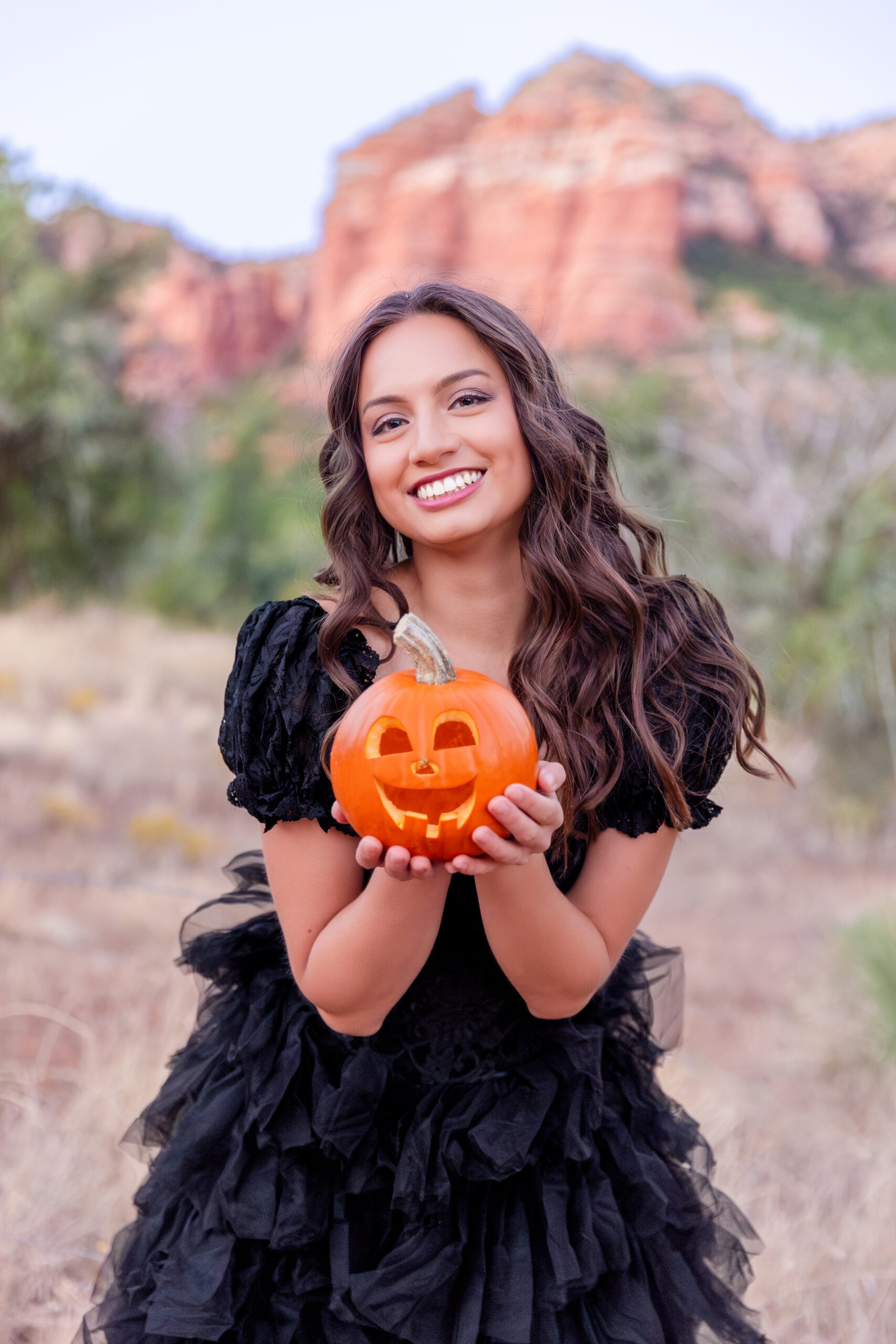 A stunning model in a flowing black gown holding a jack-o-lantern during a spellbinding Halloween sedona portrait session with Bayley Jordan Photography
