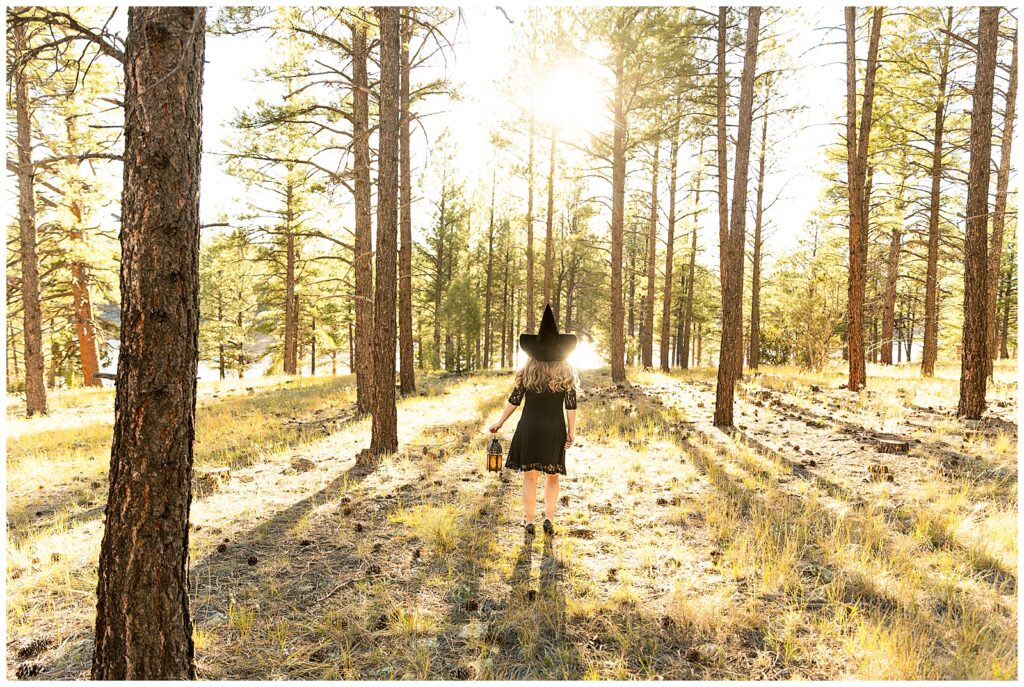 A woman stands in the woods wearing a dark pointed witch hat, lit-up lantern, and lacy black dress during a Halloween portrait session with Bayley Jordan Photography in Flagstaff