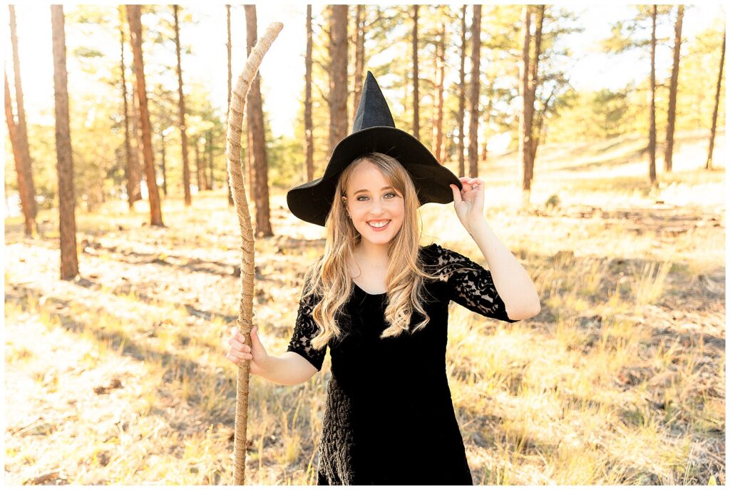 Halloween portrait session enchanted witch theme with Bayley Jordan Photography. Beautiful blonde model wears a dark black lacy gown wearing a pointed hat and leaning on a rustic broomstick.