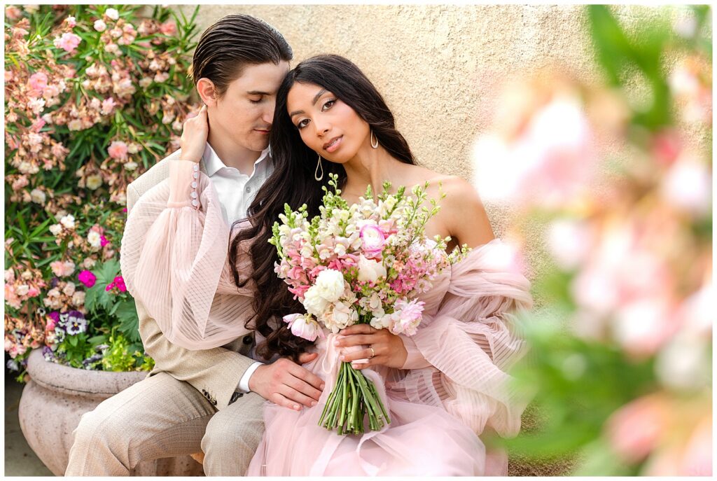 Beautiful couple surrounded by pink blossoms and holding a bundled bouquet of white and pink flowers. The couple snuggles together in high-end dress during a luxe portrait session at Tlaquepaque in Sedona, Arizona with Bayley Jordan.
